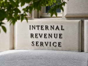 Fachada de un edificio del Internal Revenue Service con letras metálicas en la pared de piedra, parcialmente cubierta por hojas de árbol en la parte superior, representando una oficina del IRS y temas relacionados con impuestos federales.